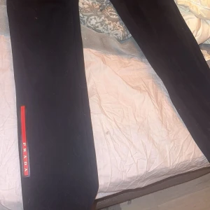 Svarta mjukisbyxor från Prada - Svarta prada mjukisbyxor med elastisk midja och dragkedjefickor. Byxorna har en röd logga längs benet och är i mjukt material, perfekta för en chill och stilren look. Jag har sytt om dem till lågmidjade för bättre passform A сору