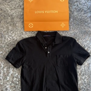 Svart pikétröja från Louis Vuitton - Svart pikétröja från Louis Vuitton med klassisk krage, tre knappar och bröstficka. Diskret rutmönster i tyget och exklusiv metallbricka med logga på sidan. Tillverkad i Italien, perfekt för dig som gillar stilrena designer.