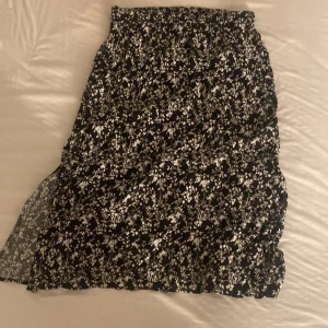 Svart blommig långkjol med slits - Svart långkjol med vitt blommönster och en snygg slits på sidan. Kjolen har en rak och flowy passform som ger en chill vibe. Perfekt för dig som gillar en romantisk och trendig stil. Resår i midjan för extra komfort.