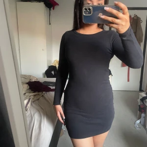 Svart tight långärmad kortklänning - Enkel och stilren svart kortklänning med lång ärm och rund halsringning. Klänningen är figurnära och har en klassisk, minimalistisk look som passar perfekt till sneakers eller boots. Mjuk och stretchig känsla för en bekväm passform. 