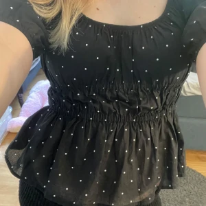 Svart prickig blus med puffärm - Supersöt svart blus med vita prickar och smockad midja som ger en snygg siluett. Blusen har korta puffärmar och lätt volang nertill. Perfekt för dig som gillar en söt och trendig stil.