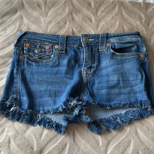 Blå jeansshorts från True Religion - Snygga blå jeansshorts från True Religion med fransig kant nertill och klassiska detaljer som kontrastsömmar och stora bakfickor med lock och knappar. Shortsen har normal passform och cool vintagekänsla, perfekta för sommaren.