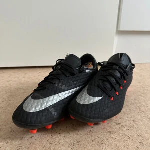 Nike Hypervenom svarta fotbollsskor - Svarta Nike Hypervenom fotbollsskor med silverfärgad swoosh och orange dobbar. Skorna har snörning och en texturerad ovandel i syntetmaterial för extra grepp på planen. Perfekta för dig som vill sticka ut med stilrena och sportiga skor på matchen.