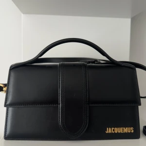 Svart Jacquemus axelväska i skinn stor modell  - Säljer en svart axelväska från Jacquemus i slätt skinn med guldiga detaljer och logga framtill. Väskan har ett stilrent, rektangulärt format med både handtag och justerbar axelrem. Invändigt är den ljus med ett innerfack. Perfekt för dig som gillar minimalistisk design.  Samt stora modellen  KVITTO FINNS
