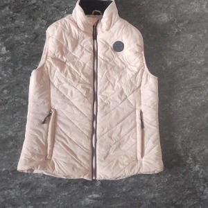 Ljusrosa quiltad dunväst från  - Supersnygg ljusrosa dunväst quiltat mönster och hög krage. Västen har två fickor med dragkedja och en diskret logga på bröstet. Perfekt att slänga över en hoodie för extra värme och stil.