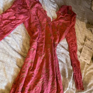 Rosa spetsklänning med v-ringning - Säljer en superfin rosa klänning i spets med v-ringad hals och långa ärmar. Klänningen har ett vackert mönster i spetsen och är figurnära upptill med lite utsvängd kjol. Perfekt för dig som vill sticka ut med en färgstark look!
