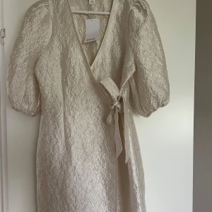 Champagnefärgad omlottklänning Monki - Superfin champagnefärgad omlottklänning från Monki med puffiga korta ärmar och knyt i midjan. Klänningen har en skimrande, lätt skrynklig yta och v-ringning. Perfekt för dig som vill sticka ut med en elegant och trendig look.