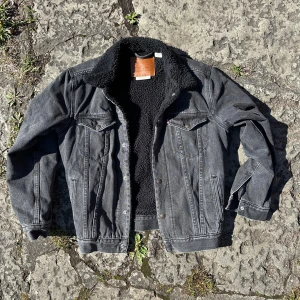 Svart sherpa jeansjacka från Levi's - Svart jeansjacka från Levi's med stor broderad logga på ryggen och mjukt svart sherpa-foder. Klassisk modell med knappar framtill, bröstfickor och krage i teddy. Perfekt för dig som vill ha en cool och varm jacka med streetkänsla.                         100% Bomull. 