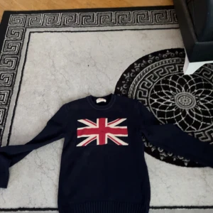 Mörkblå stickad tröja Crocker S - Mörkblå stickad tröja från Crocker i storlek S. Tröjan har ett stort Union Jack-flagga-motiv framtill i rött, vitt och blått. Tillverkad i 100% bomull med ribbade muddar och rund hals. Perfekt för dig som gillar brittisk stil.