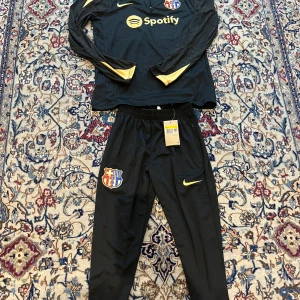 FC Barcelona Nike långärmad träningsset - Snyggt svart långärmat träningsset från Nike och FC Barcelona. Tröjan har gul Nike-logga, Spotify-tryck och klubbmärke på bröstet. Byxorna har klubbmärke, gul Nike-logga och gul detalj längs benet. Materialet är lätt och andas, perfekt för fotbollsträning.