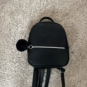 Svart ryggsäck med pälsboll - Minimalistisk svart ryggsäck i skinnimitation med justerbara axelband och ett praktiskt ytterfack med silverfärgad dragkedja. Väskan har en fluffig svart pälsboll som detalj och ett stilrent, clean look som passar till det mesta. Och har bara andvändts ett fåtal gånger!☺️