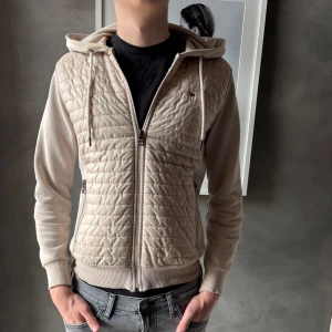 Beige quiltad jacka Tommy Hilfiger - Snygg beige quiltad jacka från Tommy Hilfiger med huva och dragkedja. Jackan har ribbade muddar, två sidofickor med dragkedja och broderad logga på bröstet. Perfekt för dig som vill ha en stilren och bekväm look.