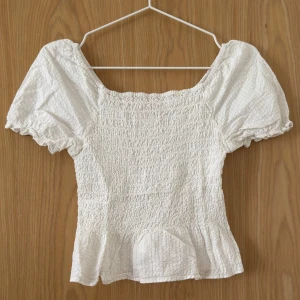 Vit blus med puffärm från Kappahl - Supersöt vit blus från Kappahl med smockad kropp och volang nertill. Blusen har korta puffärmar och en lätt v-ringning baktill. 