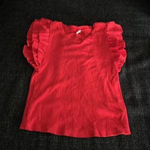 Röd volangtopp från KappAhl YOUNG - Supersöt röd topp från KappAhl YOUNG med volangdetaljer på axlarna. Modellen är ärmlös och har en rund halsringning. Tunt, ribbat tyg i bomull gör den både bekväm och snygg. Perfekt för dig som vill sticka ut med färg och volanger! Kanske passar mer till julen pga av färg men om du gillar rött så är denna tröja perfekt för dig