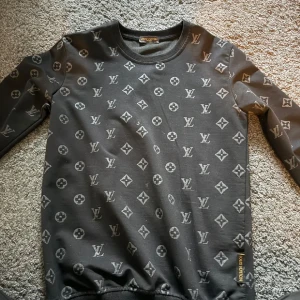 Svart Louis Vuitton tröja med monogram - Svart långärmad tröja från Louis Vuitton med ikoniskt monogrammönster över hela plagget. Rund halsringning och diskret Louis Vuitton-logga i gult på sidan. Perfekt för dig som vill sticka ut med en lyxig streetstyle-look.
