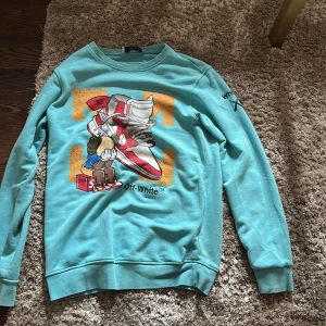 Turkos Off-White sweatshirt med print - Säljer en turkos sweatshirt från Off-White med stora grafiska prints fram och bak, bland annat sneakers och KAWS-inspirerade figurer. Tröjan har rund hals, långa ärmar och Off-White-tryck på ärmen. Tillverkad i 100% bomull för skön känsla.