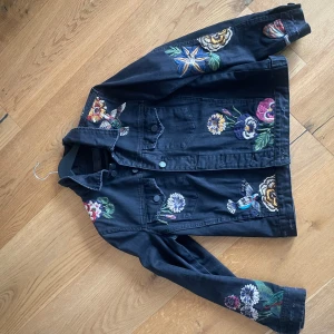 jeansjacka  - Svart jeansjacka från BlankNYC med färgglada broderade blommor och fåglar på både ärmar och framstycke. Jackan har klassisk jeansjacke-modell med knappar framtill och coola detaljer som gör den unik. Perfekt för dig som vill sticka ut lite extra.