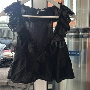 Svart volangblus med broderade detaljer M - Supersöt svart blus från Monki med volanger och broderade detaljer längs urringningen och axlarna. Ärmlös modell med luftig passform och lätt, tunn bomullskänsla. Storlek M