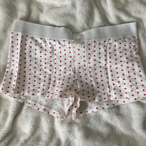 Vita shorts med röda hjärtan Gina Tricot - Supersöta vita shorts från Gina Tricot med små röda hjärtan över hela tyget. De har bred resår i midjan, tre knappar framtill och är gjorda i mjuk bomull. Perfekta för chill dagar hemma eller som pyjamasshorts.