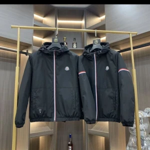 Svart vindjacka från Moncler - Svart vindjacka från Moncler med huva och dragkedja framtill. Jackan har Moncler-logga på bröstet och snygga rödvita detaljer längs dragkedjan och på ärmarna. Perfekt för dig som vill ha en stilren och sportig look.