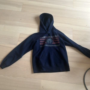 Svart Saint Laurent hoodie med tryck - Svart hoodie från Saint Laurent med vintageinspirerat tryck i rött och vitt framtill. Tröjan har huva med snörning och en stor magficka. Perfekt för dig som gillar streetstyle och vill ha något som sticker ut.