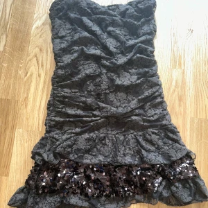 Brun spetsklänning med paljetter - Supersnygg brun kortklänning med blommig spets och volangkant nertill. Klänningen är axelbandslös och har en glittrig paljettbård längst ner som ger extra partykänsla. Perfekt för dig som vill sticka ut med en unik och elegant look.