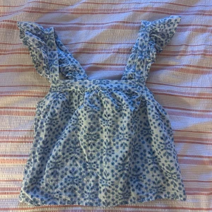 Blommig volangtopp från Pigalle XS - Supersöt topp från Pigalle i storlek XS, med blått blommönster på vit botten. Toppen har breda axelband med volangdetaljer och knytning i ryggen. Perfekt för en somrig look och riktigt skön i lätt bomullstyg.