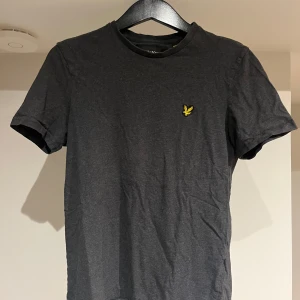 Mörkgrå t-shirt från Lyle & Scott - Mörkgrå t-shirt från Lyle & Scott med klassisk passform och korta ärmar. T-shirten har rund halsringning och en gul broderad logga på bröstet. Tillverkad i mjuk bomull som känns skön mot huden. Tröjan är i bra skick! Bara att höra av sig vid eventuella frågor!😉