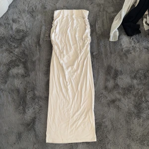 Beige långkjol - Snygg beige långkjol med rynkade detaljer längs sidorna. Kjolen har en rak passform och är tillverkad i ett mjukt, stretchigt material som ger en skön känsla. Perfekt för dig som gillar en stilren och enkel look.