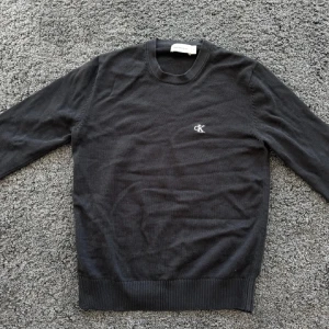 Svart Calvin Klein stickad tröja - Svart stickad tröja från Calvin Klein med diskret CK-brodyr på bröstet. Tröjan har rund halsringning, långa ärmar och ribbade muddar vid ärmslut och nederkant. Perfekt för dig som gillar stilren och enkel design.