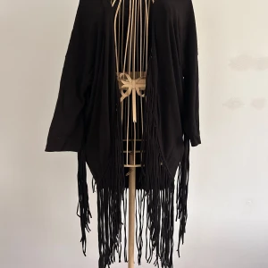 Svart kimono cardigan med fransar - Svart kimono från H&M med coola fransdetaljer längs hela fram- och baksidan. Jackan är tillverkad i mjuk bomull med lite stretch för extra komfort. Perfekt för dig som vill sticka ut med en unik och trendig look. Stl S