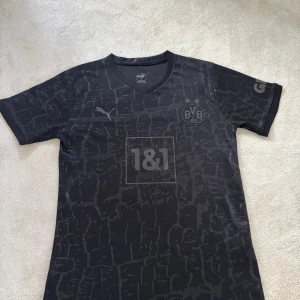 BVB Dortmund Reus svart matchtröja Puma - Snygg svart Borussia Dortmund fotbollströja från Puma med Reus 11 på ryggen. Tröjan har korta ärmar, V-ringning och ett unikt mönster i mörkgrått. BVB-logga och Puma-märke på bröstet, samt sponsortryck framtill. Tillverkad i lätt och ventilerande material.