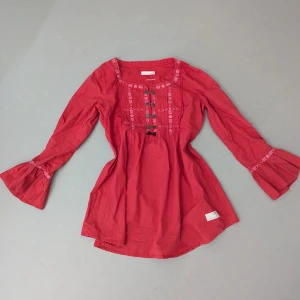 Röd broderad blus Odd Molly - Supersöt röd blus från Odd Molly med broderade detaljer framtill och utsvängda långa ärmar. Blusen har en rak passform, rundad halsringning och knappar framtill. Perfekt för dig som gillar boho-vibbar och färg!