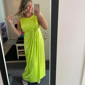 Limegrön plisserad långklänning - Säljer en limegrön långklänning med plisserat tyg och ärmlös design. Klänningen har en snygg detalj med drapering och glittrig brosch i midjan. Perfekt för dig som vill sticka ut med färg och stil på nästa event!helt ny