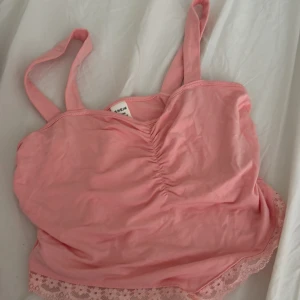 Rosa linnetopp med spets från SHEIN - Supersöt rosa linnetopp från SHEIN med smala axelband och rynkdetalj framtill. Toppen har en fin spetskant nertill med blommönster som ger en extra gullig touch. Perfekt för varma dagar och enkel att matcha med jeans eller shorts.