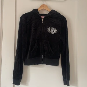 Svart velourjacka Juicy Couture  - Svart velourjacka från Juicy Couture med huva och dragkedja. Ikoniskt broderat emblem i vitt på bröstet och stor logga på ryggen. Mjuk känsla, ribbade muddar och kängurufickor framtill. Perfekt för en chill och trendig look. Använt ett fåtal gånger. 
