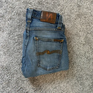 Blå jeans från Nudie Jeans, storlek 28/32 - Säljer ett par klassiska blå jeans från Nudie Jeans med snygga slitningar och kontrastsömmar. Modellen har fem fickor och Nudie-loggan på bakfickan. Jeansen är i bomull och har en rak passform som funkar till det mesta.