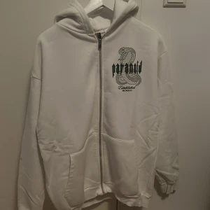 Vit hoodie Paranoid med ormtryck - Vit hoodie från Paranoid med stor orm och textprint både fram och bak. Dragkedja framtill, två fickor och huva. Oversized passform och långa ärmar, perfekt för en streetwear-look.