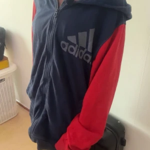 Adidas hoodiejacka i blått och rött - Mysig hoodiejacka från Adidas med marinblå kropp och röda ärmar. Stor Adidas-logga på bröstet, dragkedja framtill och huva. Perfekt för chill dagar eller när du vill ha en sportig look.