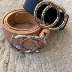 Två bälten med dubbelring-spänne - Säljer två snygga bälten med trendigt dubbelring-spänne i silver. Ett bälte är svart och det andra är beige, båda i skinnimitation. Svarta bältet är 75 cm och det beige är 65 cm. Perfekta accessoarer för att lyfta din outfit.