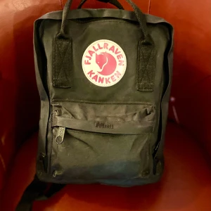 Svart Fjällräven Kånken ryggsäck - Klassisk svart Fjällräven Kånken ryggsäck med ikonisk rund logga framtill i rött och vitt. Ryggsäcken har ett stort huvudfack, framficka med dragkedja och två sidofickor. Tillverkad i slitstark syntet med justerbara axelremmar och bärhandtag upptill.