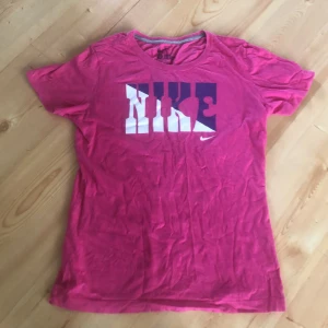 Rosa Nike t-shirt med tryck, slim fit - Snygg rosa t-shirt från Nike i slim fit-modell. Stor logga i vitt och lila framtill. Klassisk rund halsringning och korta ärmar. Tillverkad i mjuk bomull, perfekt för en sportig och avslappnad look. Känns som medium. Ska stryka den om du köper den. Den är från 2011.