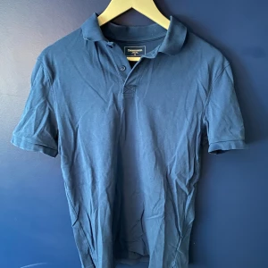 Blå pikétröja från Dressmann M - Klassisk blå pikétröja från Dressmann i storlek M. Tröjan har korta ärmar, krage och knäppning med två knappar framtill. Tillverkad i mjuk bomull som känns skön mot huden. Perfekt för en clean och enkel look.