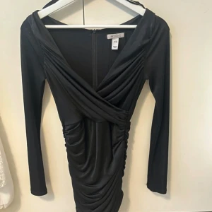 Svart omlottklänning från NLY One - Supersnygg svart kortklänning från NLY One med omlott-detalj och draperad effekt framtill. Klänningen har lång ärm, v-ringning och dragkedja i ryggen. Perfekt för dig som gillar en tight och stilren look.