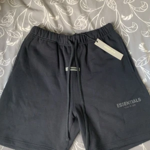 Svarta shorts Essentials Fear of God - Svarta shorts från Essentials Fear of God med elastisk midja och snörning. Klassisk loose fit med en bakficka och logga framtill. Mjukt bomullsmaterial som är perfekt för chill dagar eller träning. Snygg minimalistisk design och riktigt skön passform.