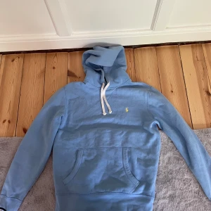 Ljusblå hoodie från Polo Ralph Lauren - Snygg ljusblå hoodie från Polo Ralph Lauren med klassisk gul logga på bröstet. Tröjan har huva med vita snören och en stor magficka. Perfekt för dig som gillar en clean och stilren look. Mjuk bomullskvalitet och långärmad modell.