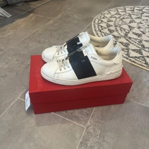 Valentino Garavani Open sneakers vit/mörkblå - Snygga Valentino Garavani Open sneakers i vitt skinn med bred mörkblå rem över mitten. Det är 100% äkta och man får med allt på som är med på bilderna. Klassisk rund tå, platt sula och coola nitar bak på hälen. Grå snörning och diskret logga på plösen. Perfekta för dig som gillar lyxiga och stilrena sneakers. Priset är inte hugget i sten!!!