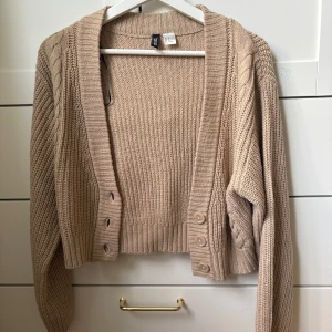 Beige stickad kofta - Supermysig beige/ljusbrun stickad kofta från H&M i croppad modell. Koftan har ribbade muddar, kabelstickade detaljer längs ärmarna och tre knappar framtill. Perfekt att slänga över en topp för en chill look. Långärmad och i mjukt material.