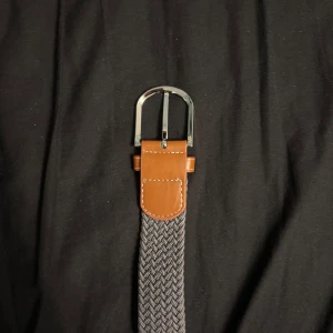 Grått flätat bälte med bruna skinn-detaljer - Snyggt grått bälte med flätad struktur och bruna skinn-detaljer vid spännet. Spännet är i silverfärgad metall och bältet har en modern, stilren look som passar till jeans eller chinos. Perfekt accessoar för att lyfta din outfit.