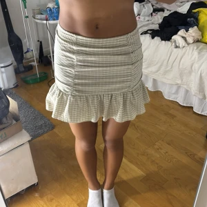 Beige kortkjol med volang från Gina Tricot - Supersöt beige kortkjol från Gina Tricot i storlek XXS. Kjolen har tunna vita ränder, smickrande rynkade detaljer framtill och en volangkant nedtill som ger en extra feminin touch. Perfekt för dig som gillar en söt och trendig look.